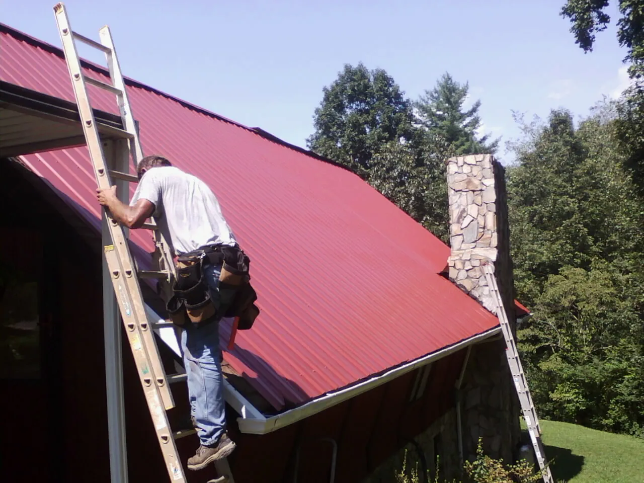 Roofer on a ladder beside a red metal roof and stone chimney — Soffit & Fascia Repair by Roofing Brothers in Astatula